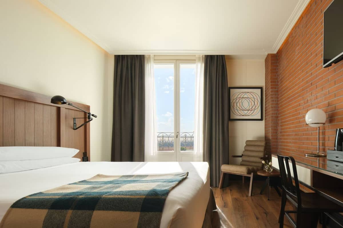 A hotel room with a bed, a desk, a chair, a TV, and a large window with curtains.