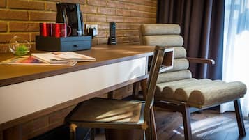 A hotel room with a brick wall, a wooden desk with a coffee maker, a remote control, and a chair with a woven seat.