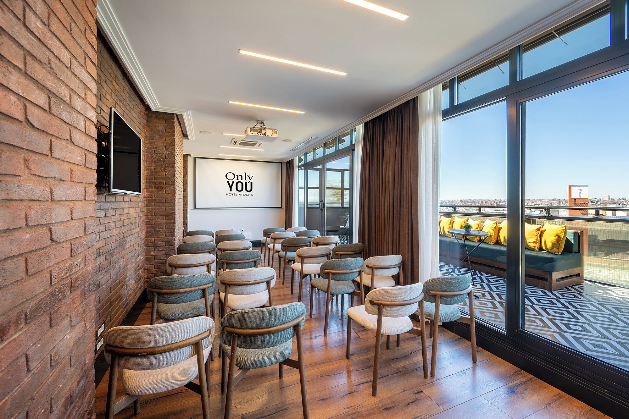 A conference room with rows of chairs, a large screen displaying 'Only YOU HOTEL ATOCHA', and a view of the city through large windows.