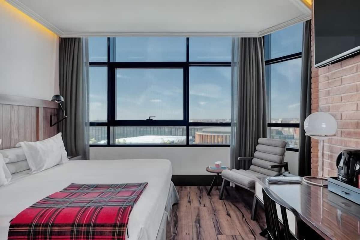 A hotel room with a bed, a desk, a chair, a window with a view, and a brick wall.