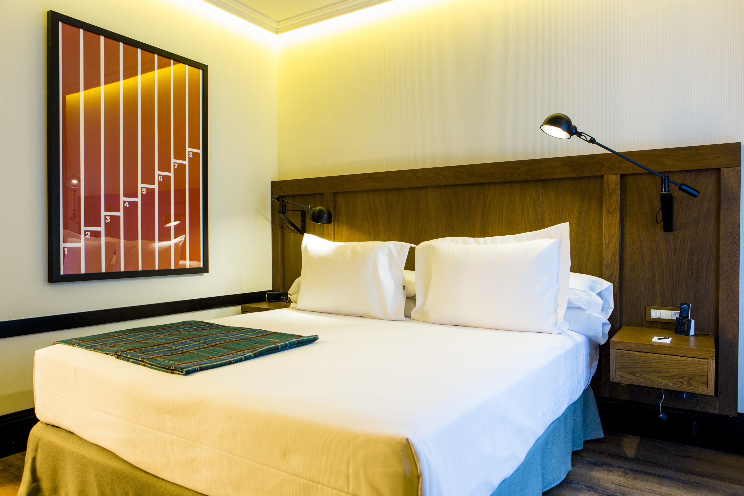 A hotel room with a large bed, a nightstand with a lamp, a wooden headboard, and a wall-mounted artwork depicting stairs.