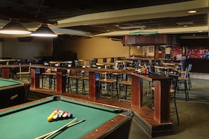 A pool table with balls and cues, surrounded by tables and chairs in a room with a high ceiling and wooden accents.