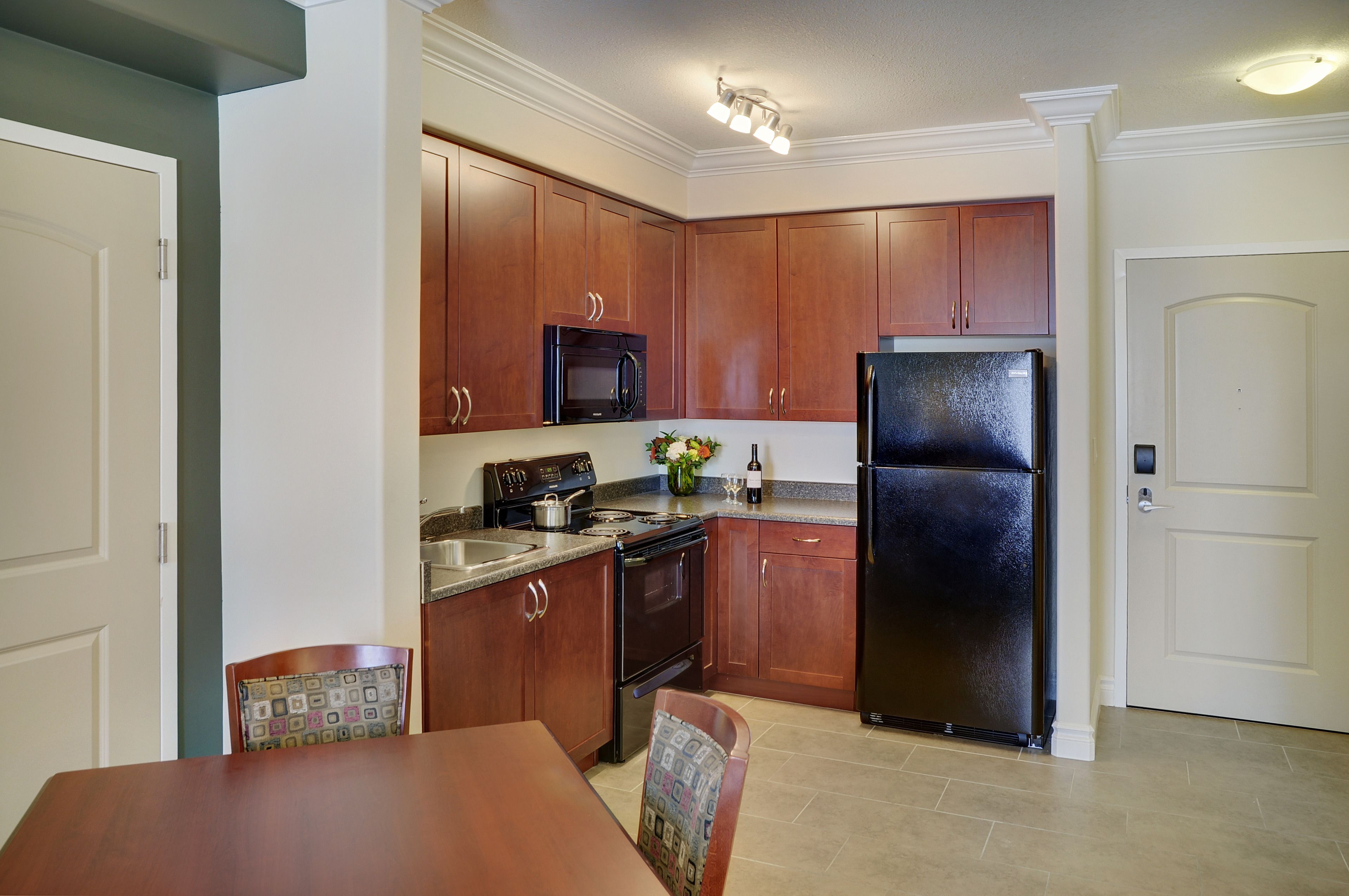 A kitchen with wooden cabinets, a black refrigerator, a stove, and a microwave. There is a dining table with chairs and a vase with flowers on the counter.