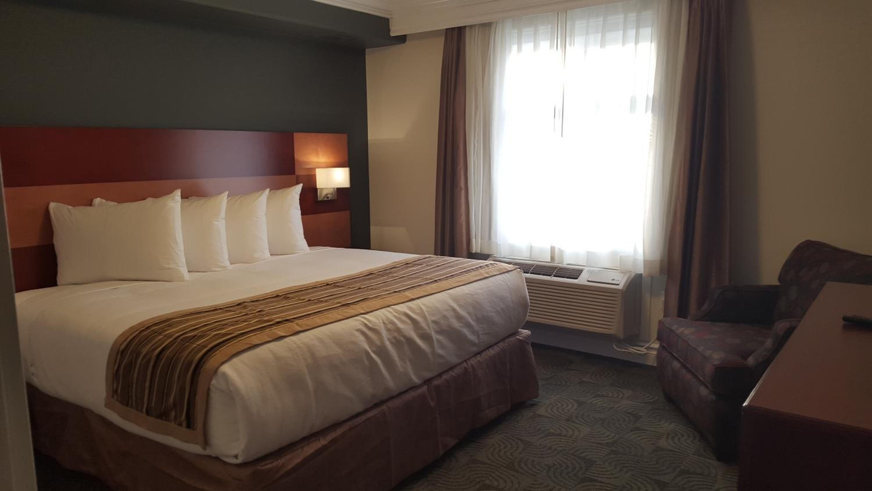 A hotel room with a large bed, a desk, a chair, a window with curtains, and an air conditioning unit.