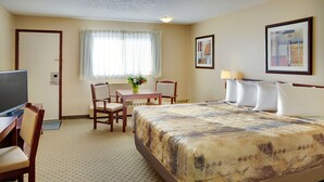 Quarto de hotel com uma cama, uma escrivaninha com cadeira, televisão, janela com cortinas e um quadro na parede.