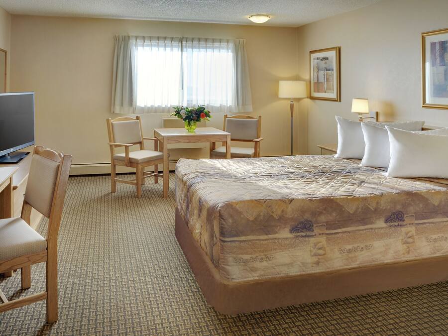 Hotelkamer met een bed, een bureau met een computer, twee stoelen, een tafeltje met een vaas en een raam met gordijnen.