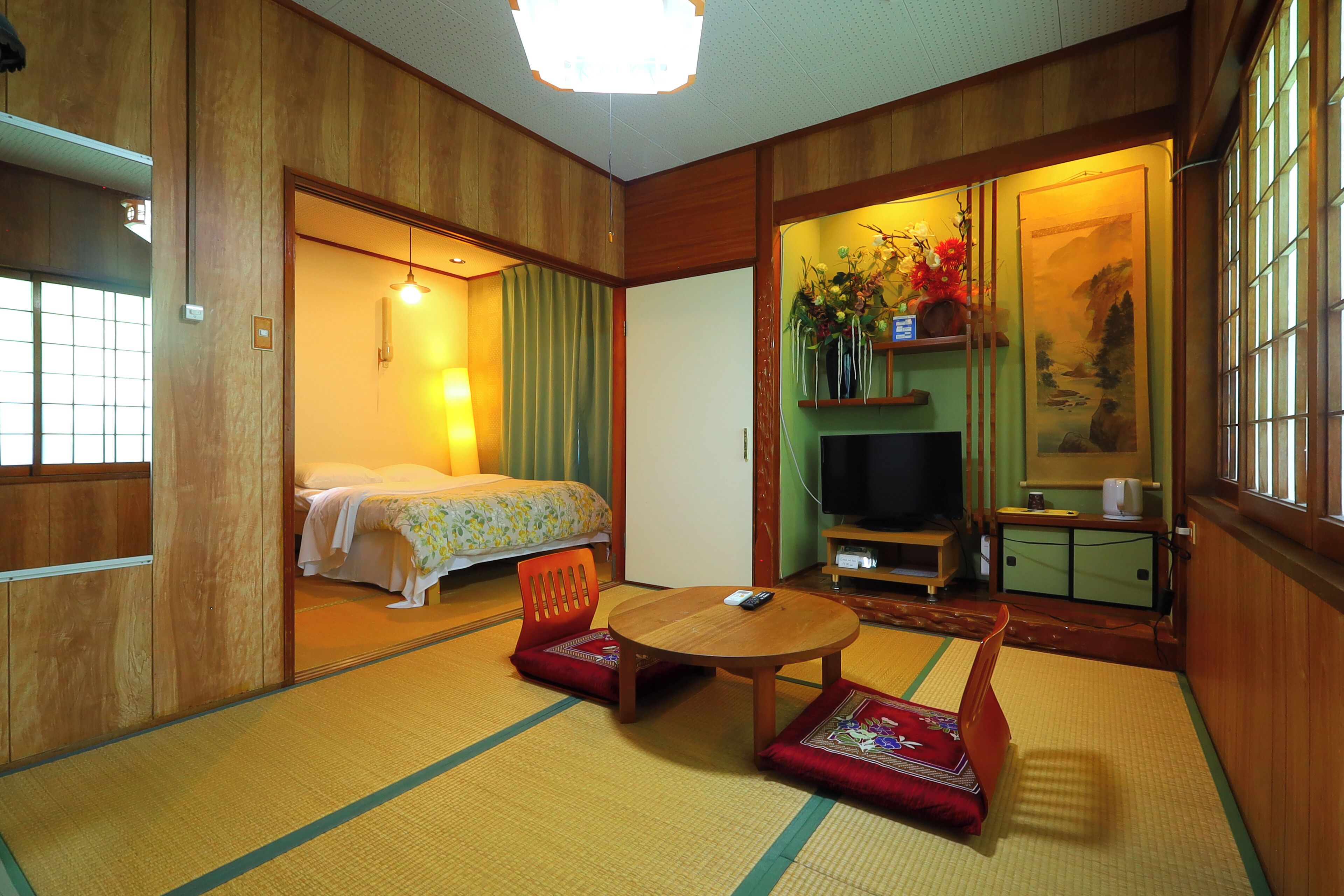 A traditional Japanese-style room with tatami flooring, a wooden bed, a television, and a small table with a red cushion.