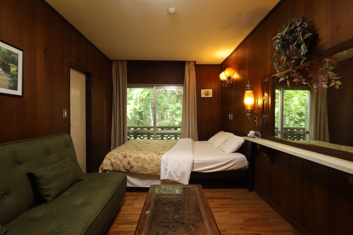 Una habitación con cama, sofá verde, piso de madera, ventana con vista a follaje y una lámpara fijada a la pared.