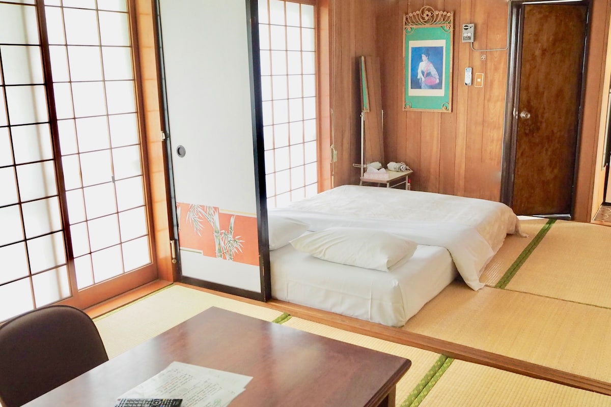 Una habitación japonesa tradicional con suelo de tatami, una cama con ropa de cama blanca, un escritorio con control remoto y un cuadro en la pared.