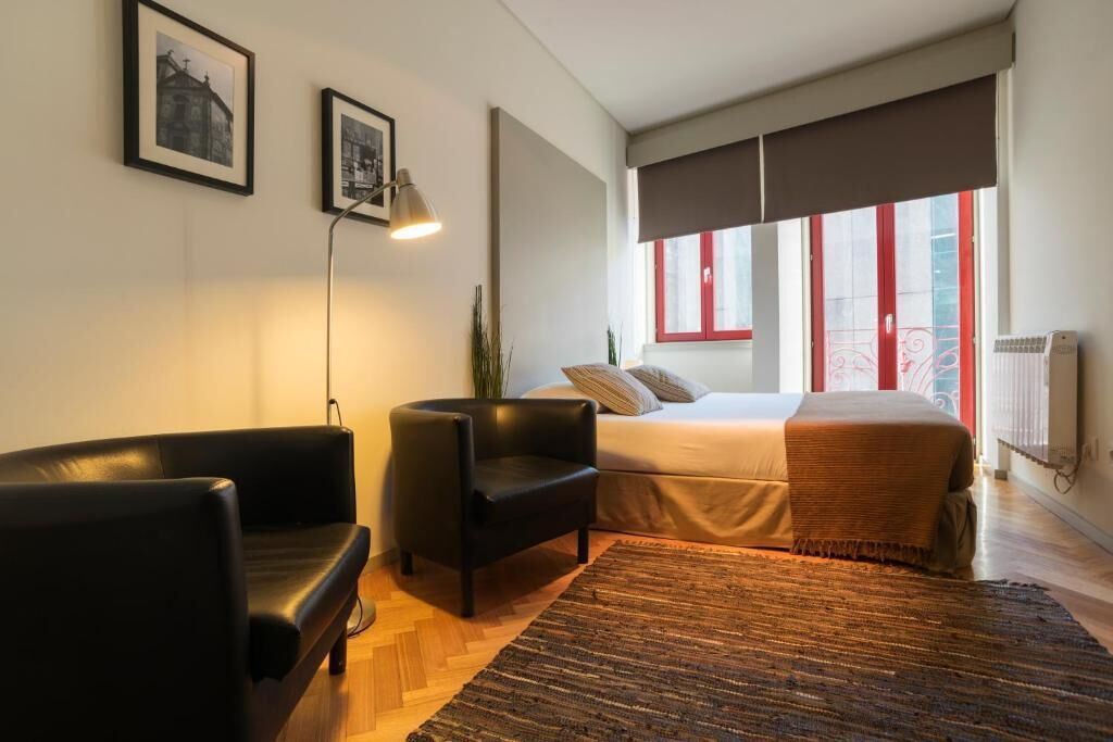 A modern bedroom with a bed, a black leather chair, a standing lamp, and a large window with red frames.