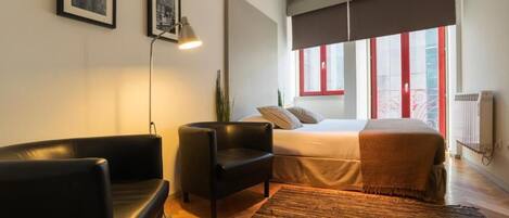 A modern bedroom with a bed, a black leather chair, a standing lamp, and a large window with red frames.