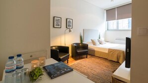 A modern hotel room with a bed, two chairs, a desk with water bottles, a lamp, and framed pictures on the wall.