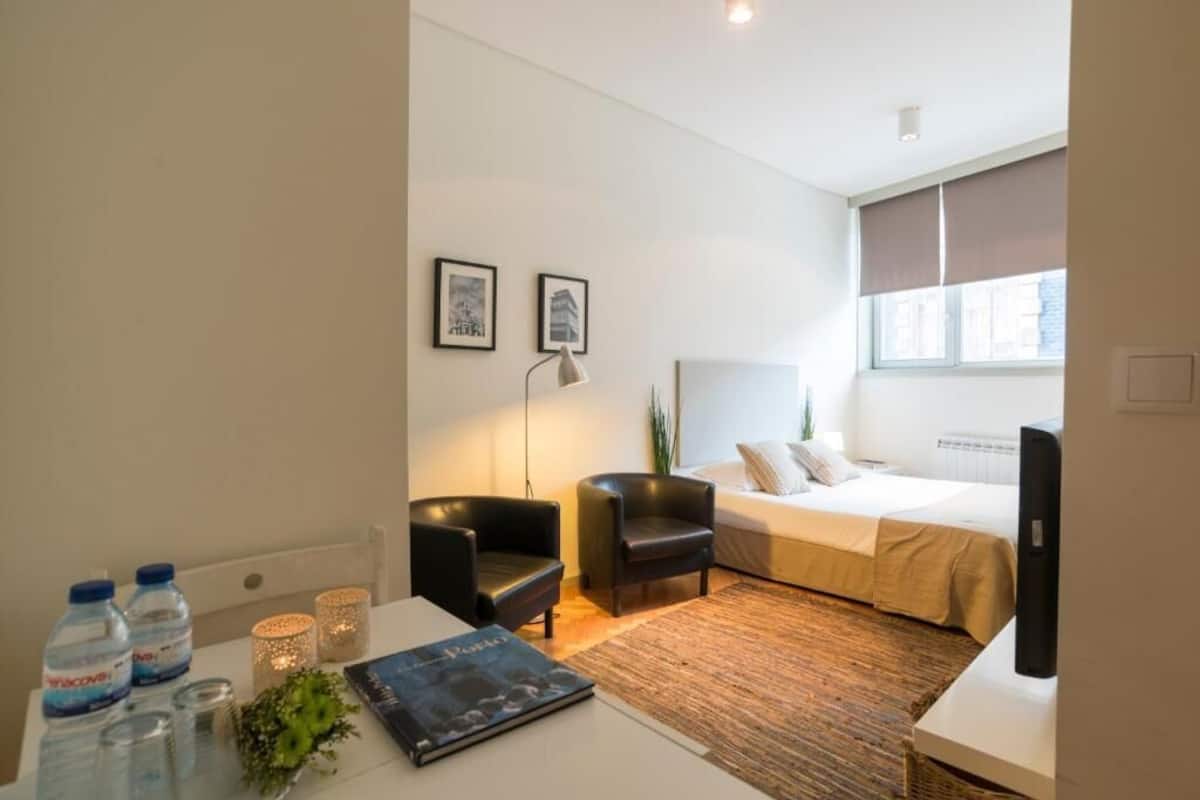 A modern hotel room with a bed, two chairs, a desk with water bottles, a lamp, and framed pictures on the wall.