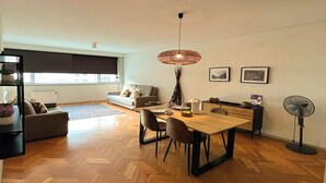 A spacious living area with a wooden herringbone floor, a dining table with chairs, a sofa, and a standing fan.