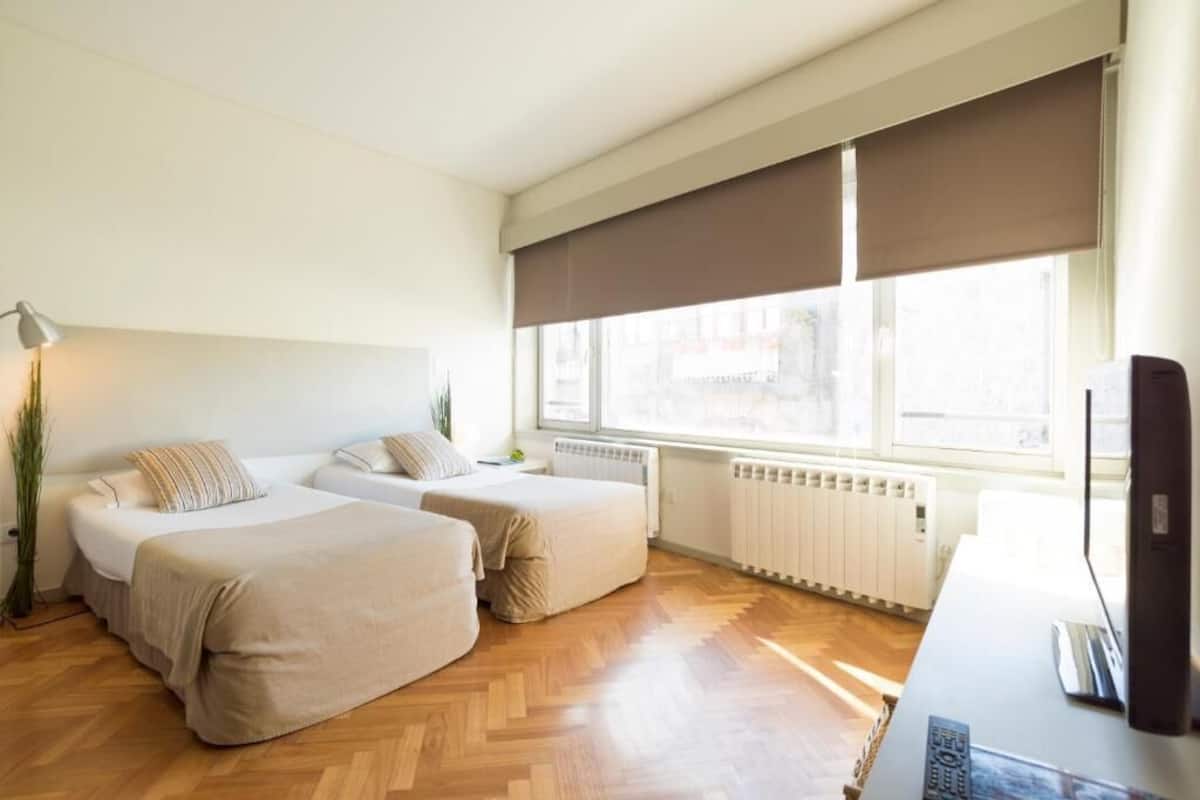A hotel room with two beds, a desk with a TV, and large windows with blinds.