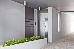 Un pasillo moderno con piso de concreto, una maceta blanca con plantas verdes y una puerta de ascensor de vidrio.