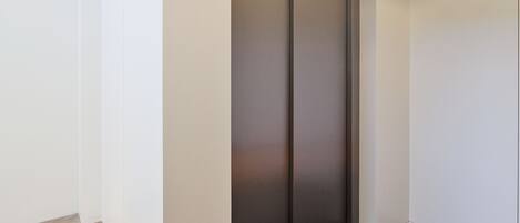 Egy modern lift egy olyan helyiségben, melynek fehér falai és csempézett padlója van.