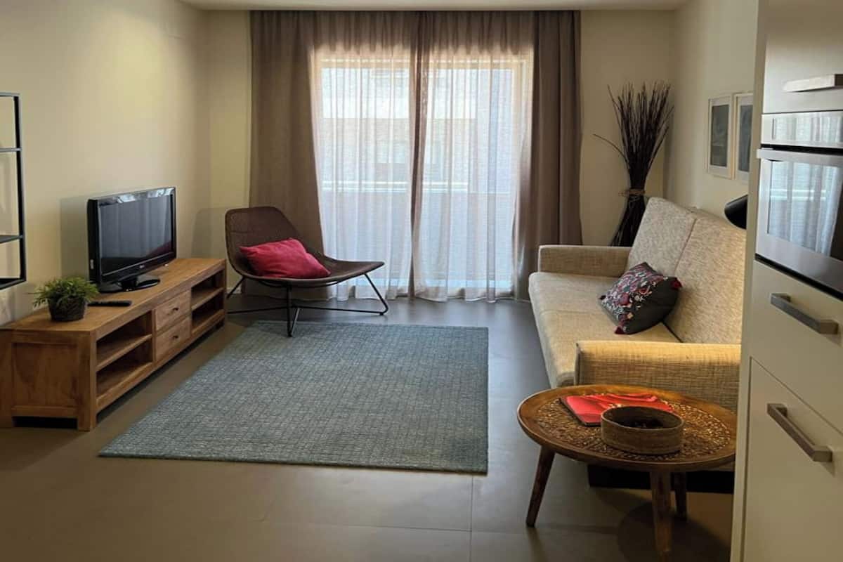 A modern living room with a flat-screen TV, a brown chair, a beige sofa, a small round table, and a large window with curtains.