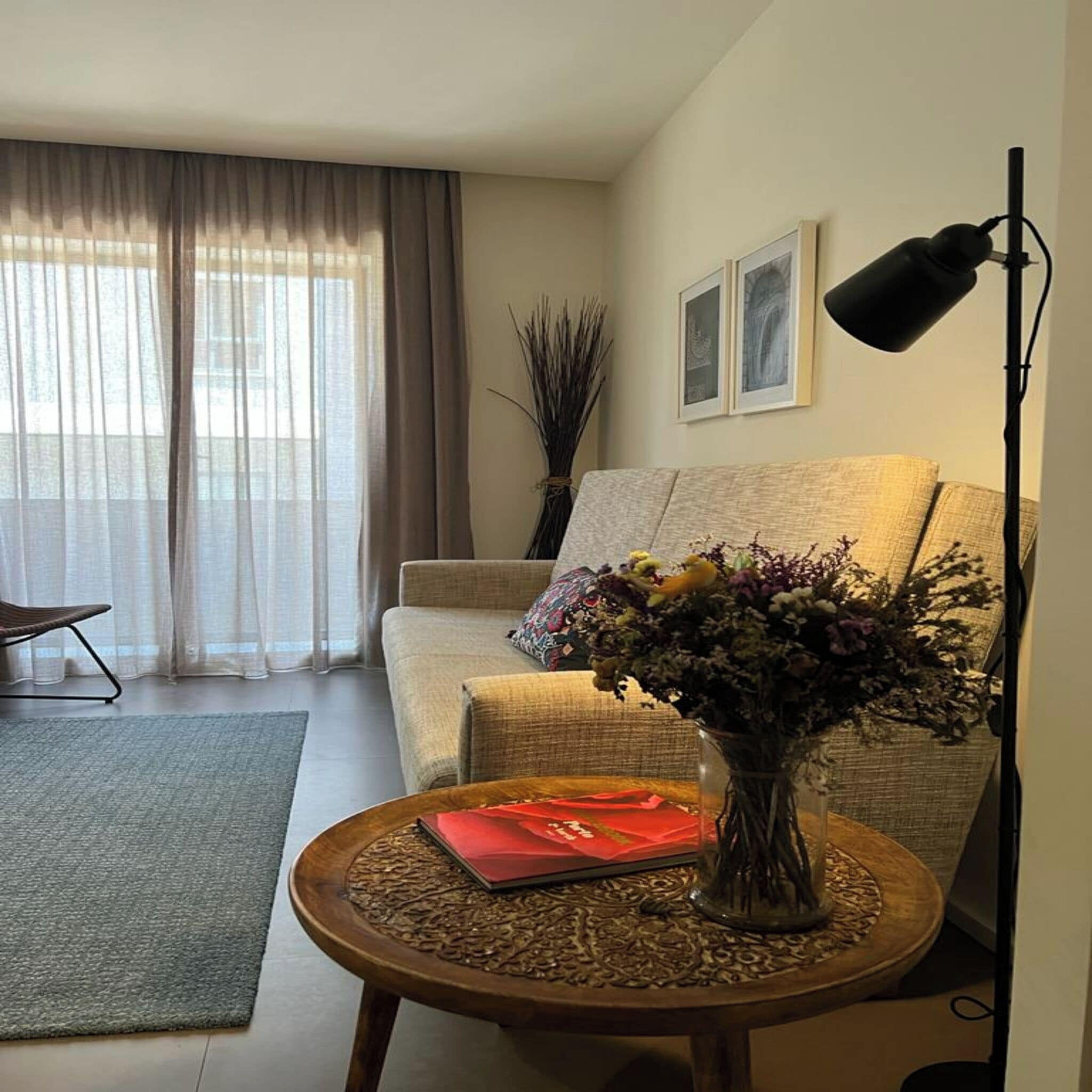 A living room with a sofa, a round wooden table with a vase of flowers, a lamp, and a chair.
