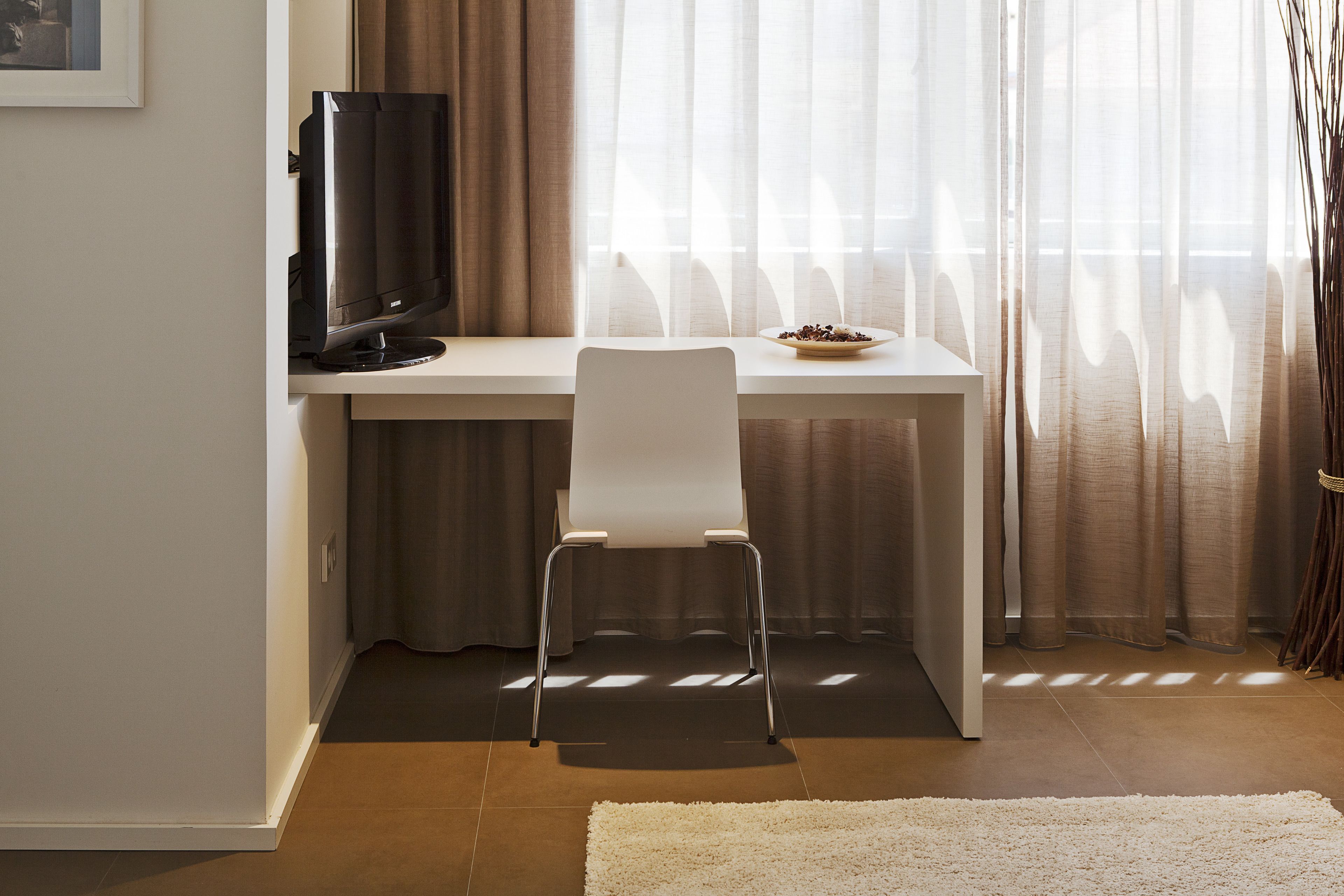 A modern room with a white desk, a chair, a television, and a window with sheer curtains.