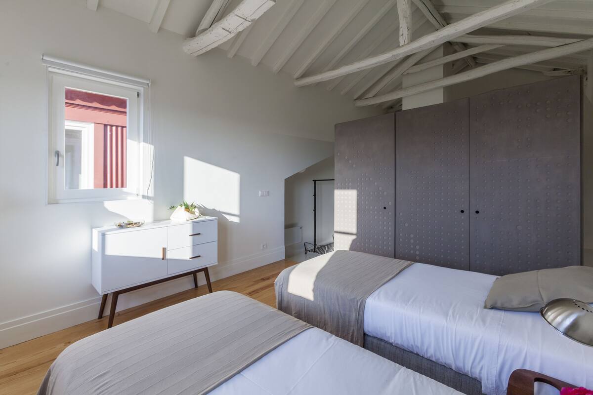 Un dormitorio con dos camas, un aparador blanco, un armario grande y una ventana con vista a un edificio rojo.