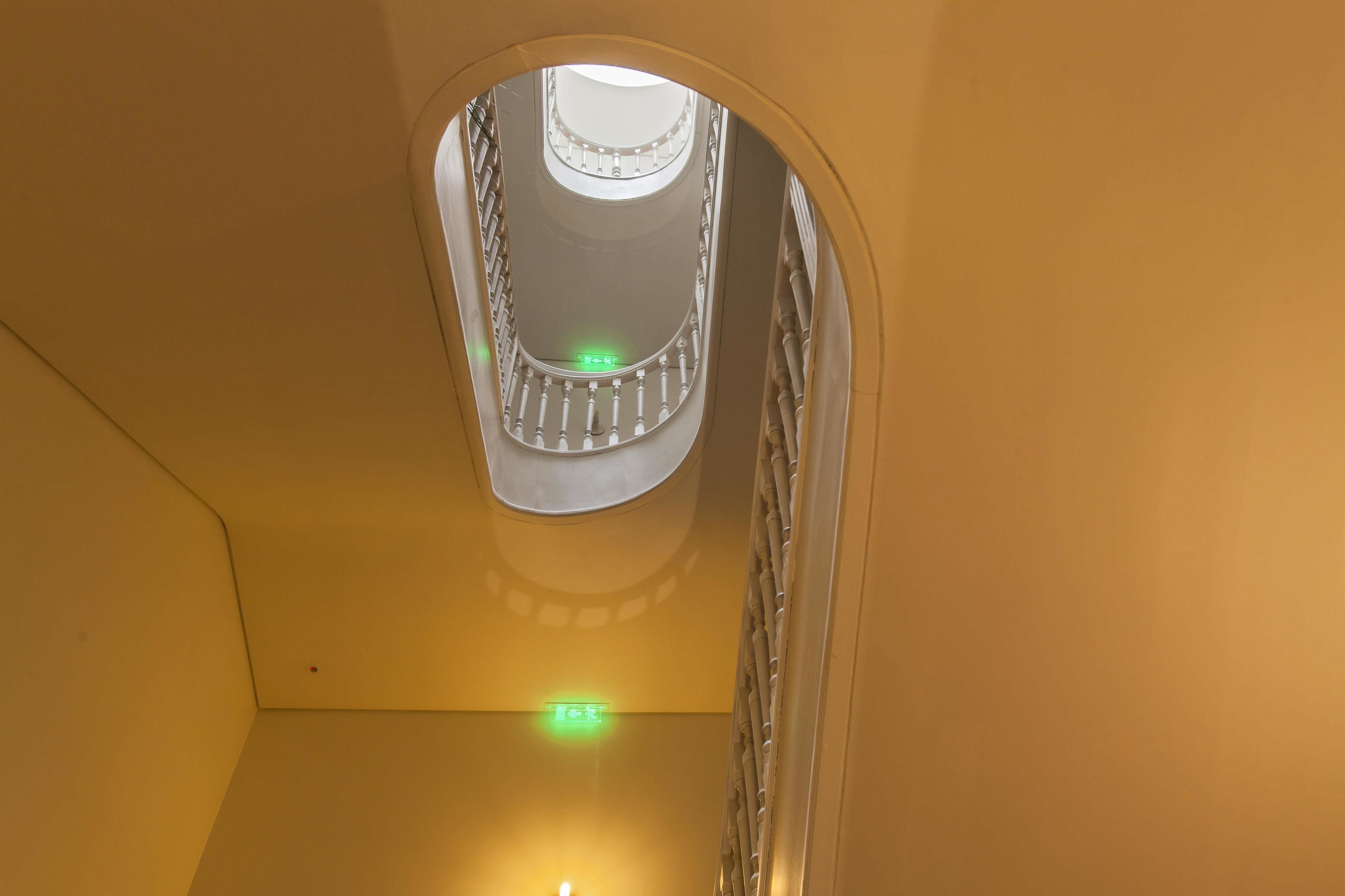 Een uitzicht op een trap met een glazen omheining, een plafondlamp en een groen vluchtroutebord.