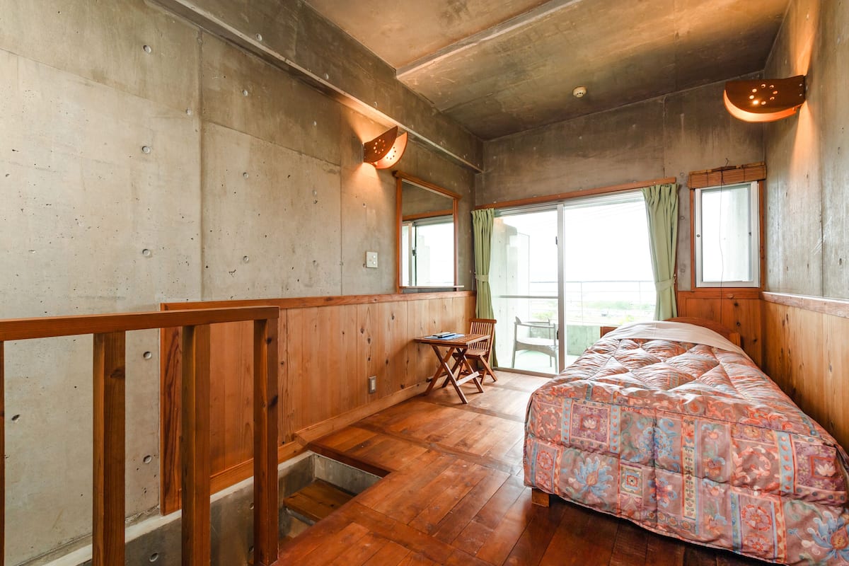 A bedroom with a wooden floor, a bed with a patterned bedspread, a wooden desk, a chair, and a window with a curtain.