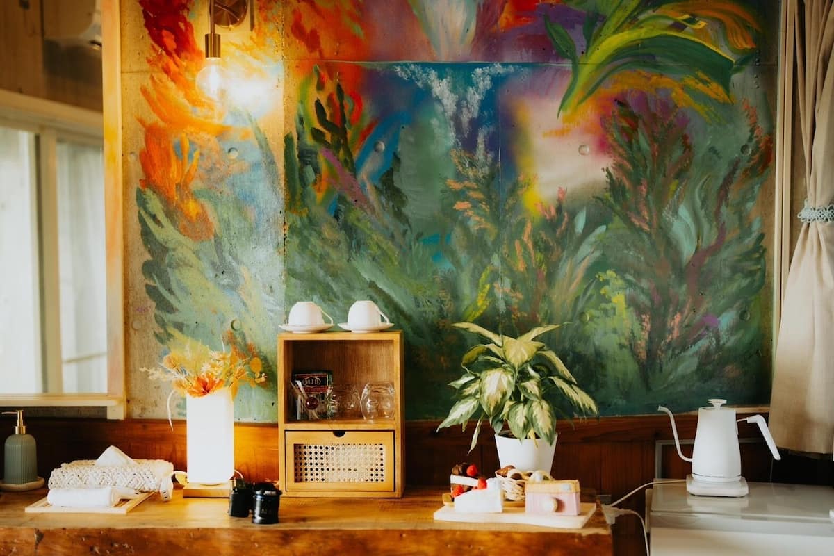 A wooden table with a white teapot, cups, and a potted plant. A colorful abstract painting is on the wall behind.