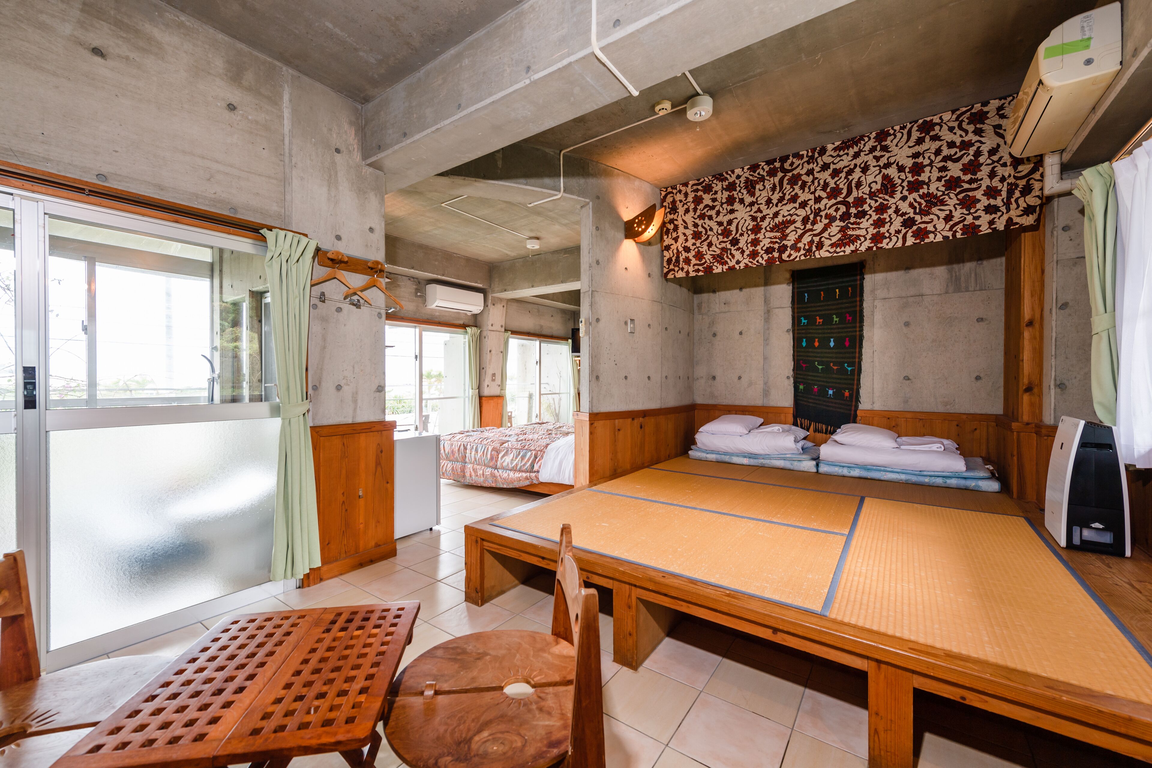 Une pièce avec un grand lit sur tatami, une table en bois et une chaise. Il y a une fenêtre avec un rideau et un climatiseur fixé au mur.