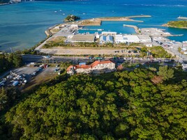 An aerial view of a coastal area with a marina, a large industrial building, and a forested hill.