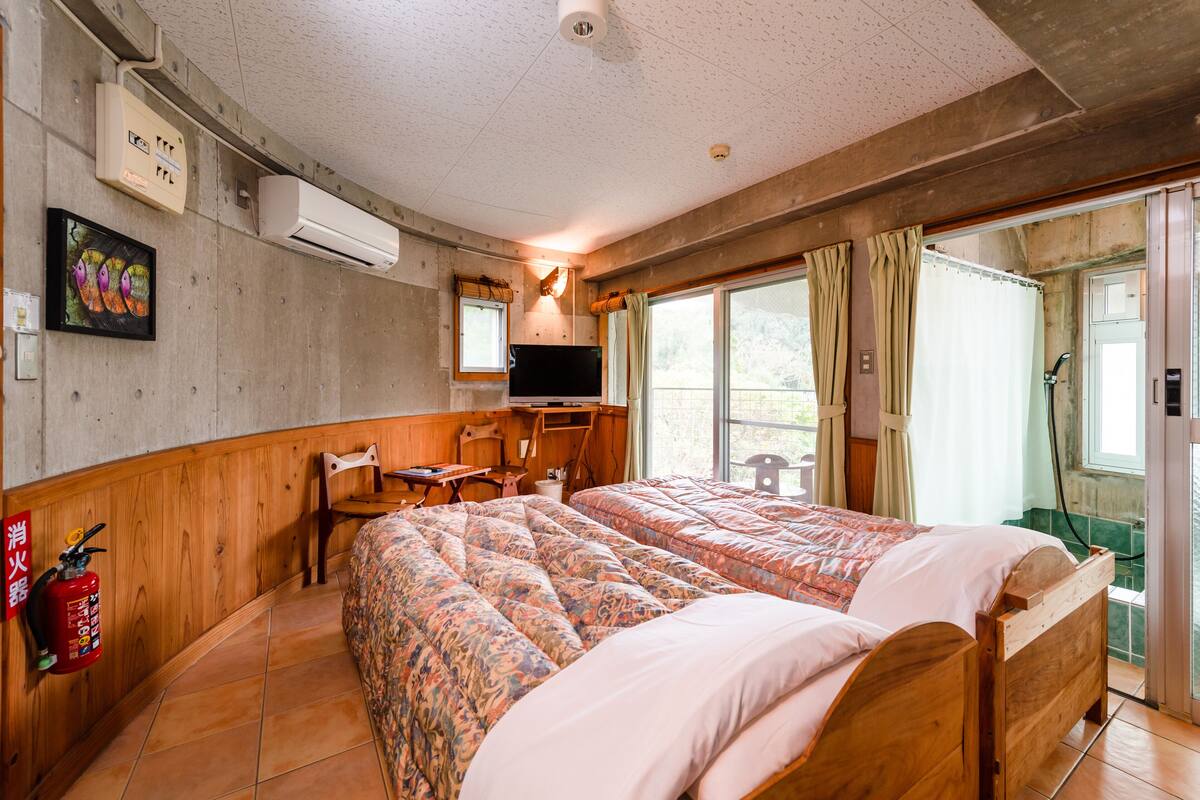 A room with two beds, a TV, a wooden desk, a chair, a fire extinguisher, and a window with curtains.