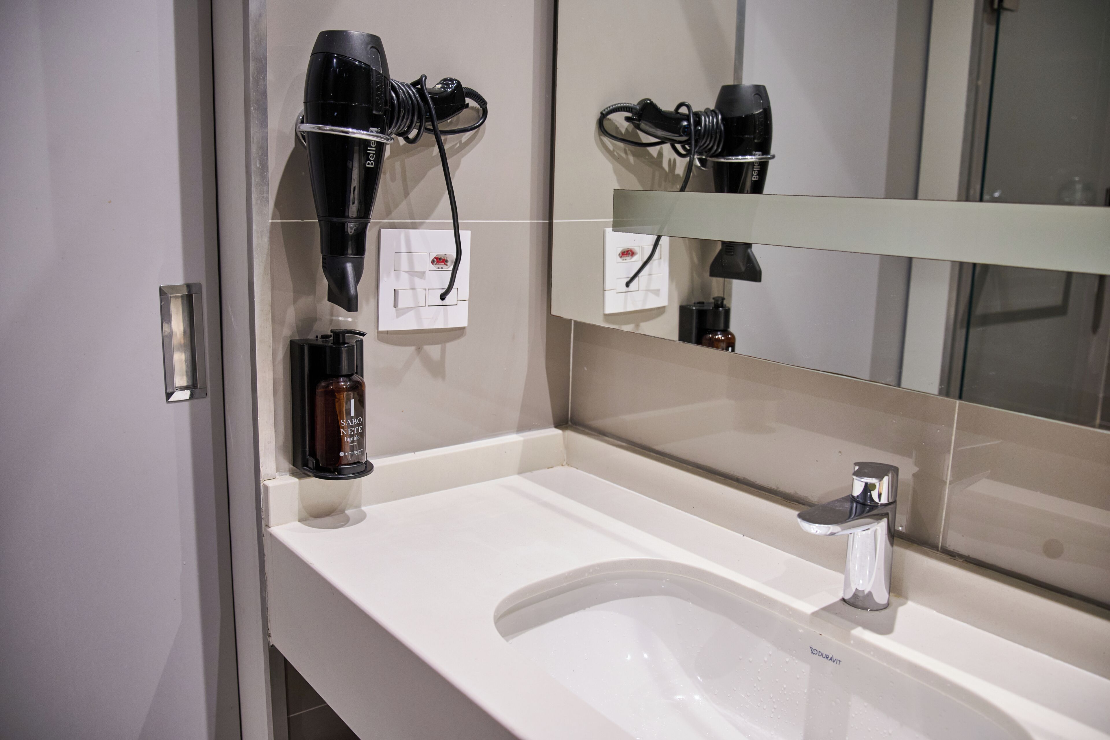 Ein Badezimmer mit Waschbecken, Spiegel, Fön und einer Flasche Shampoo.
