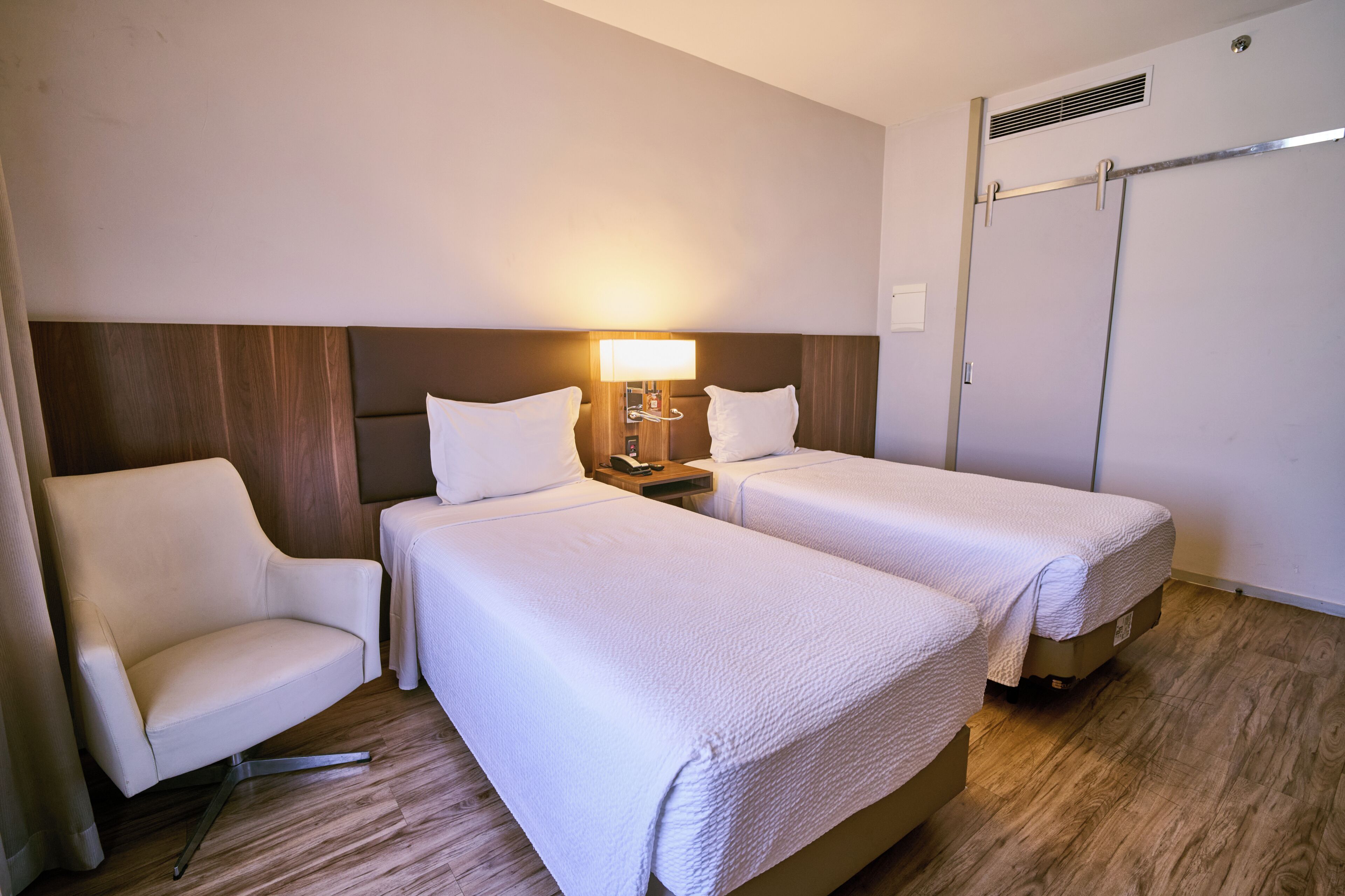 A hotel room with two beds, a chair, a nightstand with a lamp, and a wardrobe.