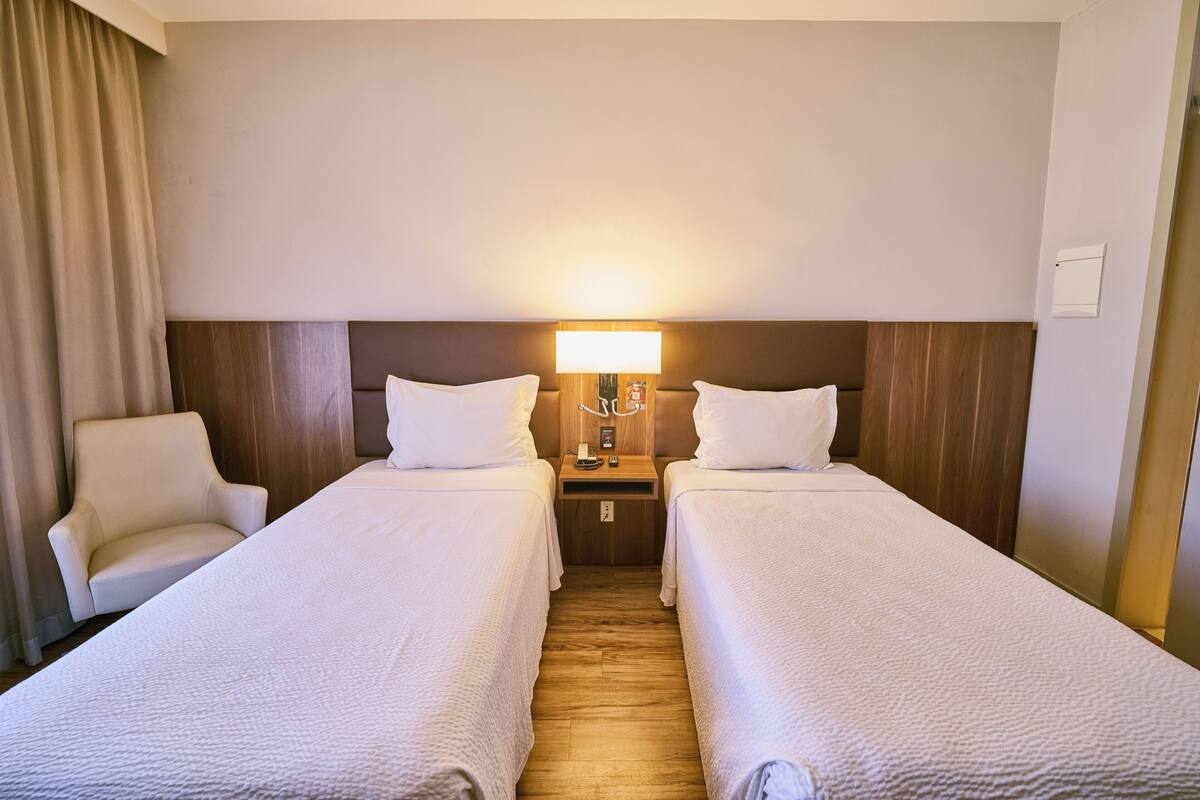 A hotel room with two beds, a nightstand with a lamp, a chair, and a wooden floor.