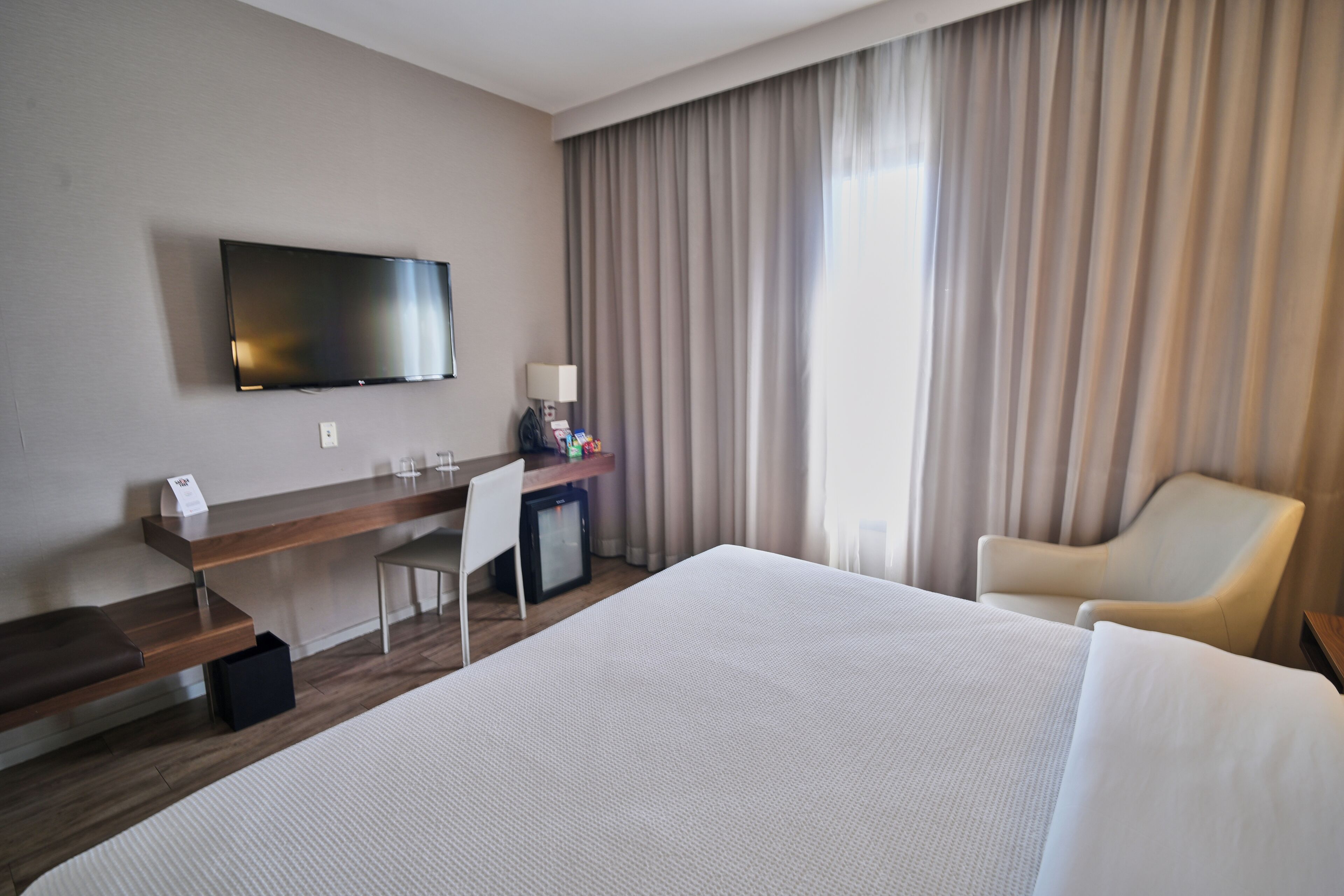 Habitación de hotel con cama, escritorio con computadora, televisor, silla y ventana con cortinas.