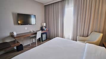 Habitación de hotel con cama, escritorio con computadora, televisor, silla y ventana con cortinas.
