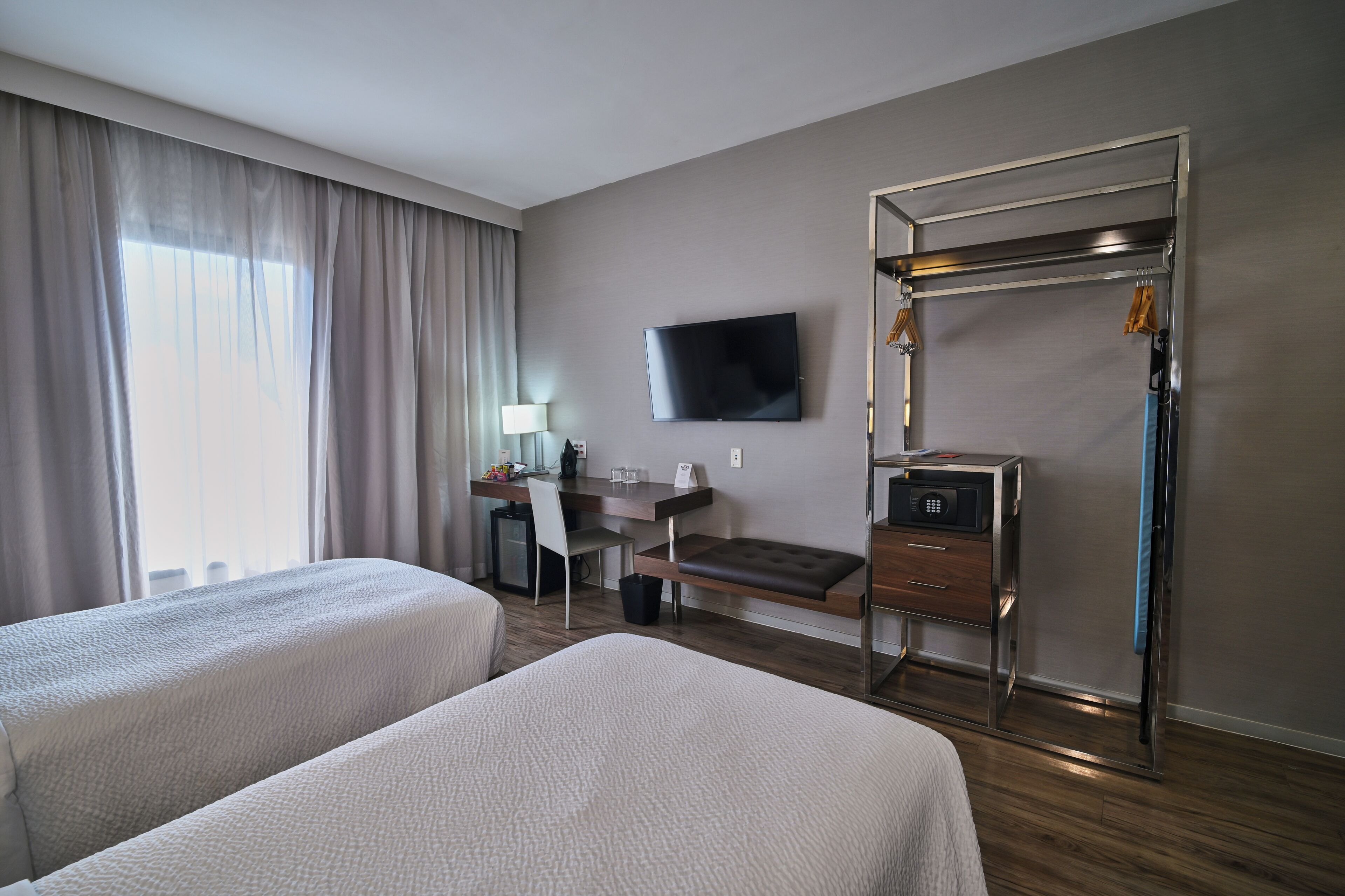Habitación de hotel con dos camas, un escritorio con silla, televisor, perchero y caja fuerte.