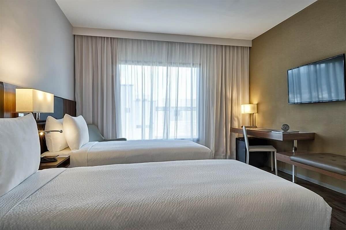 A hotel room with two beds, a desk, a chair, a TV, and a large window with curtains.