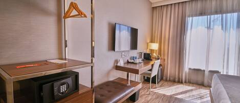 Habitación de hotel con escritorio de madera, televisor, banca, perchero y ventana con cortinas.
