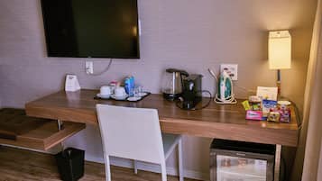 A hotel room with a flat-screen TV, a coffee maker, a mini fridge, a desk with a chair, and a small ironing board.