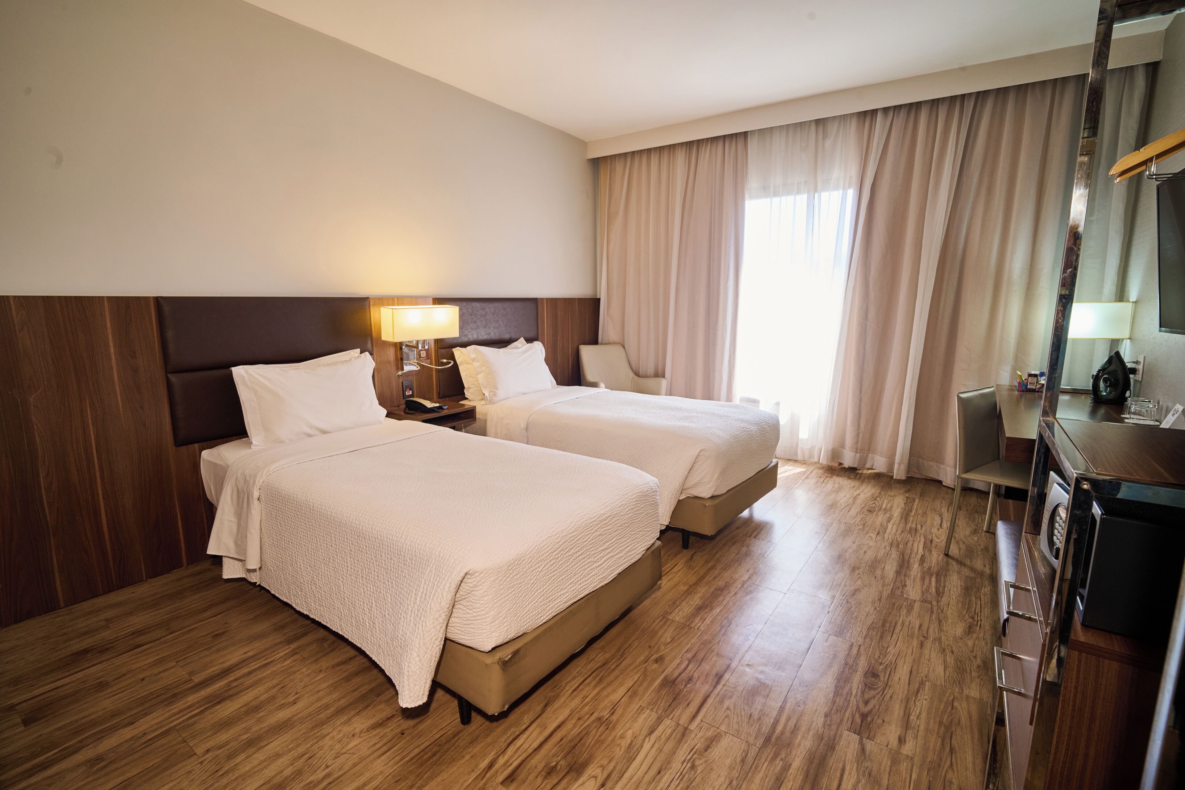 Quarto de hotel com duas camas, piso de madeira, escrivaninha com abajur e uma janela grande com cortinas.