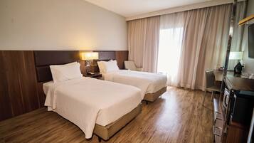 Quarto de hotel com duas camas, piso de madeira, escrivaninha com abajur e uma janela grande com cortinas.