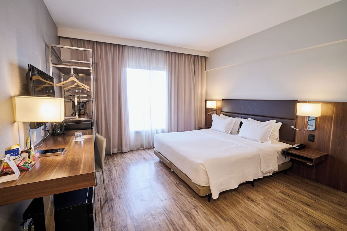 A hotel room with a bed, bedside tables, a desk with a lamp, a mirror, and a wardrobe.