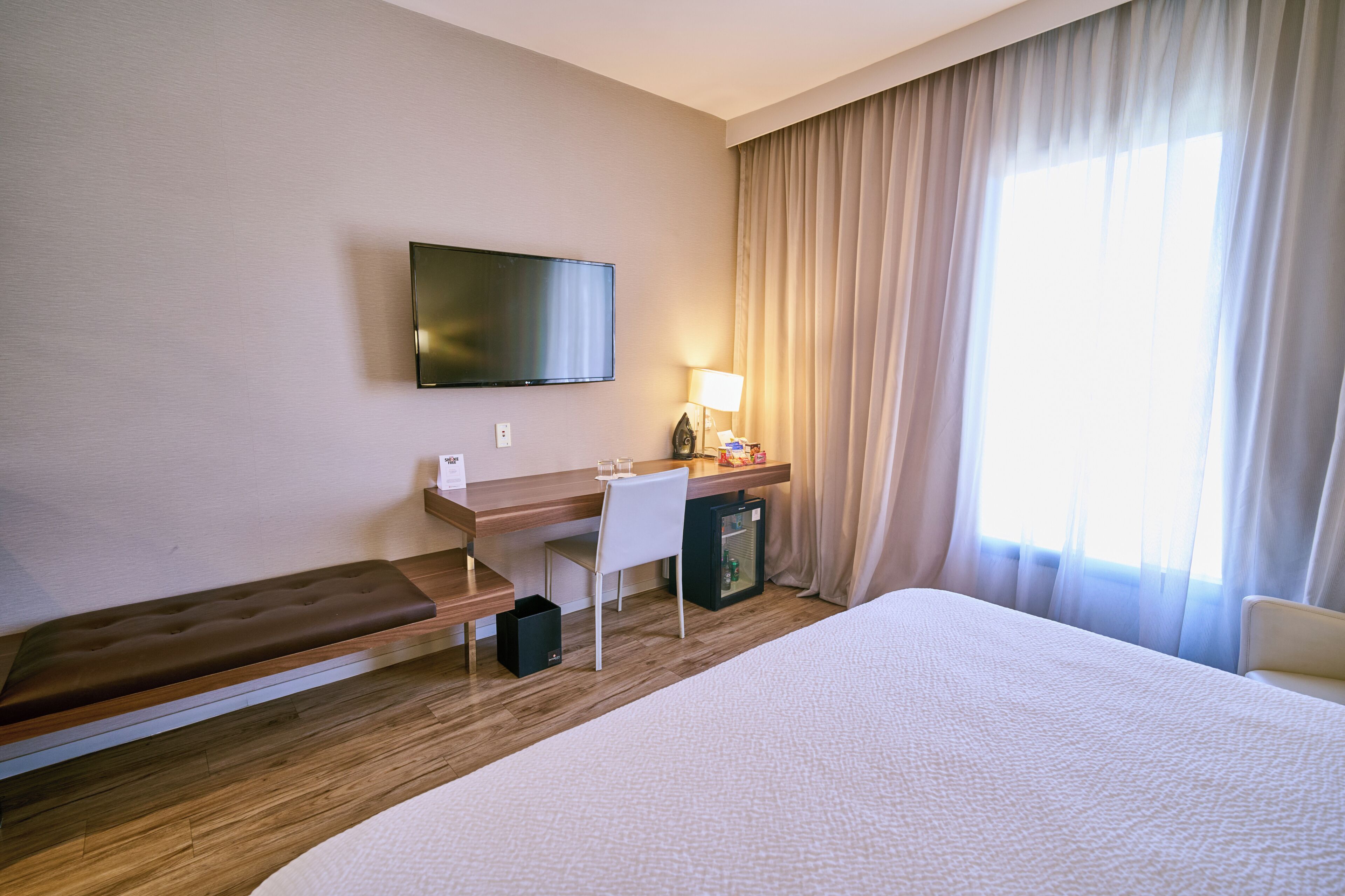 A hotel room with a bed, a desk with a chair, a television, a lamp, and a window with curtains.