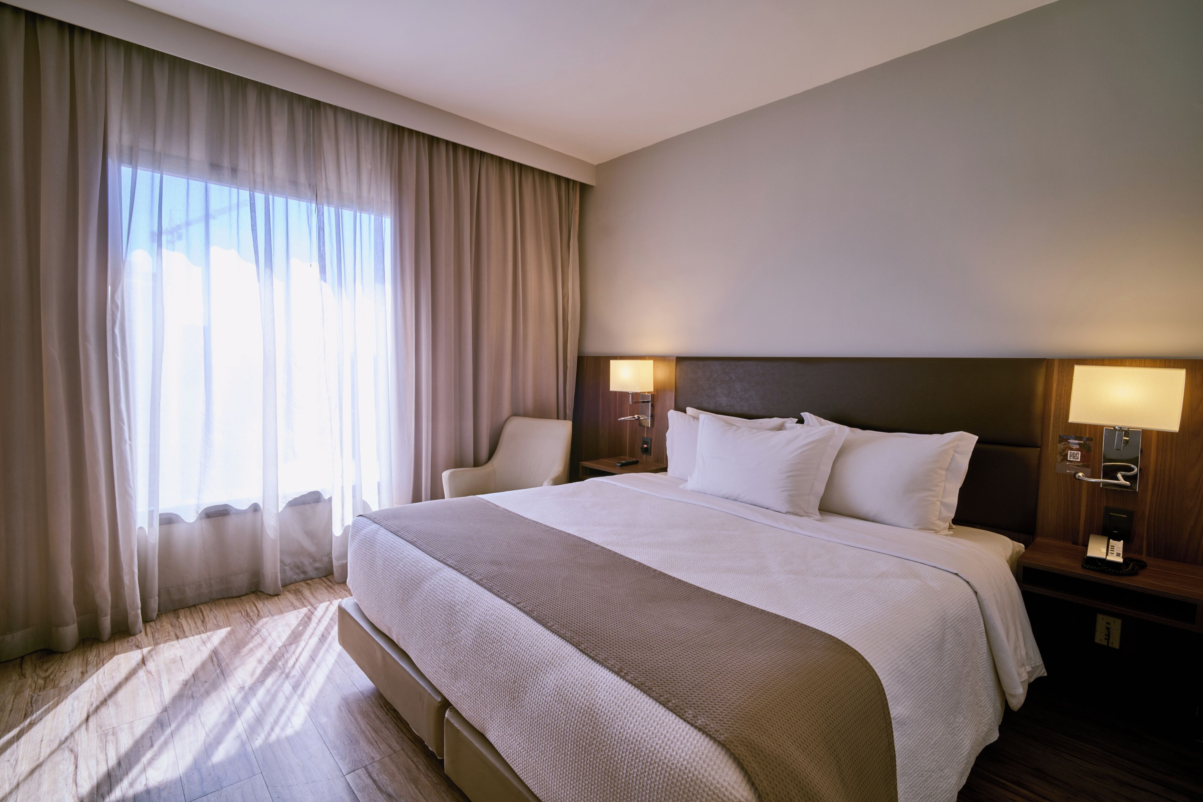 A hotel room with a large bed, two bedside lamps, a chair, and a window with curtains.