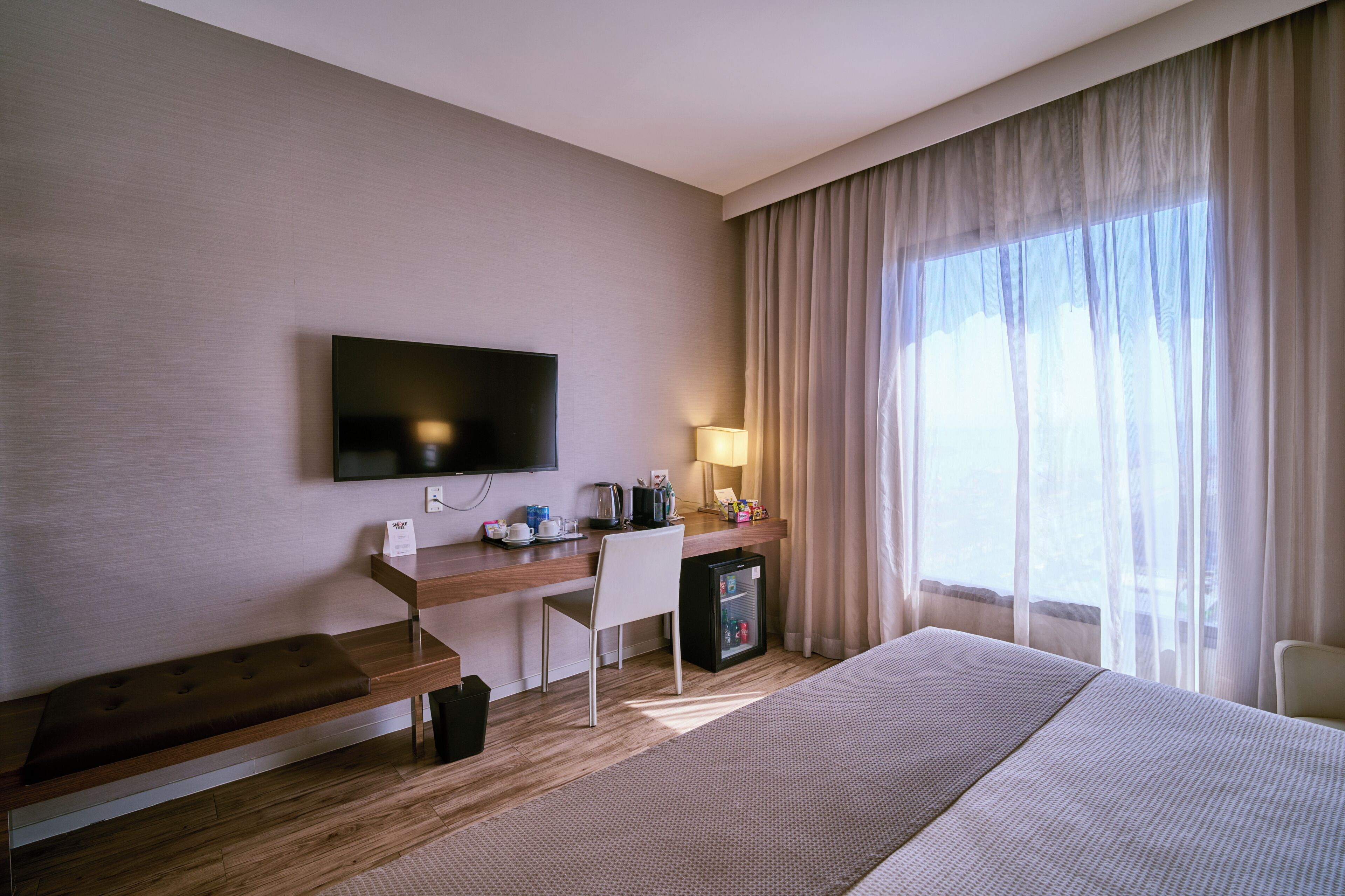 Habitación de hotel con cama, escritorio con televisor, una silla, refrigerador pequeño y ventana con cortinas.