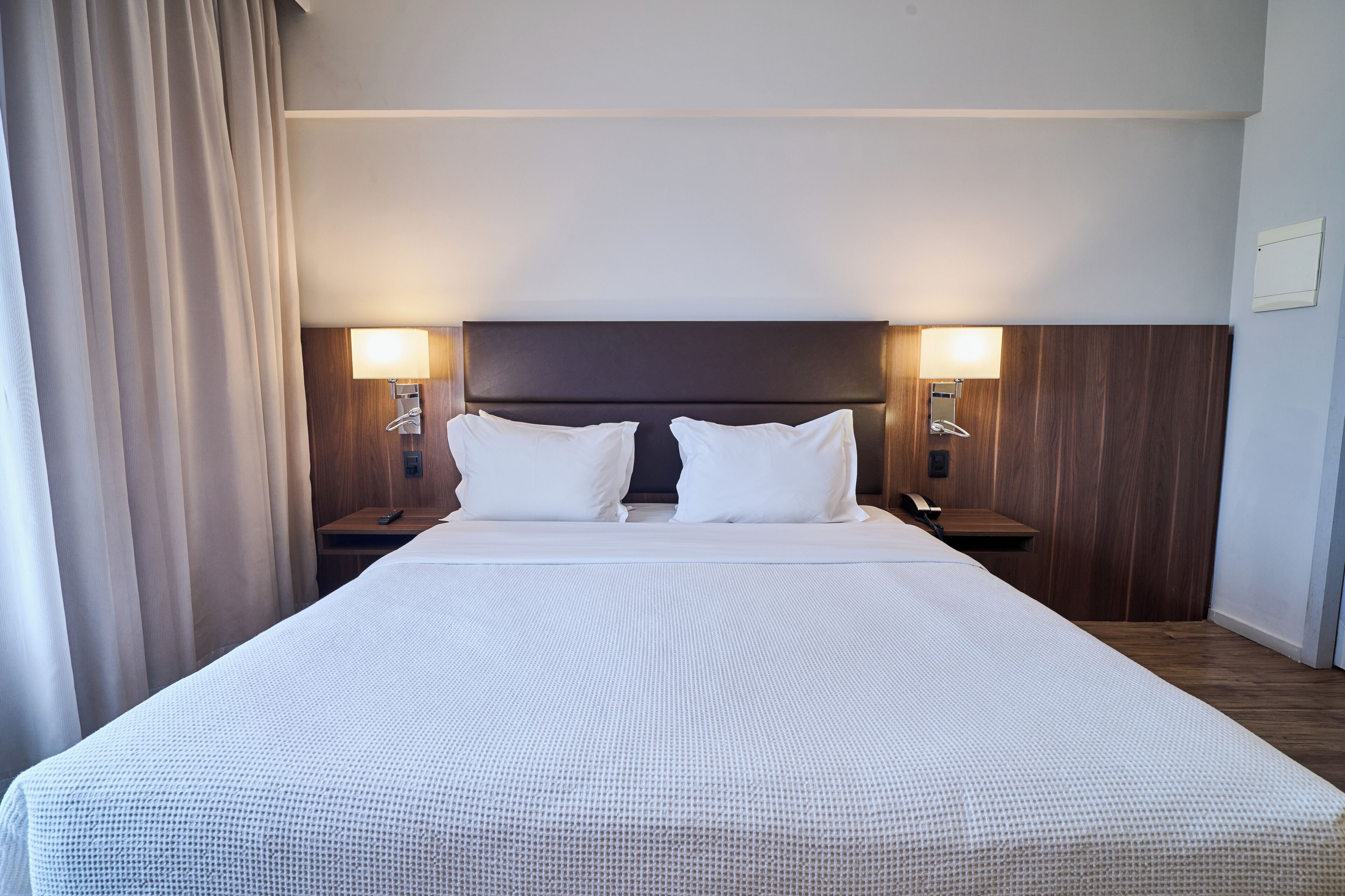 A hotel room with a large bed, two bedside tables, a wooden headboard, and two wall-mounted lamps.