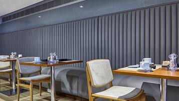 Un café moderne avec des tables et des chaises en bois, un banc recouvert d’un tissu gris et un mur à motifs.