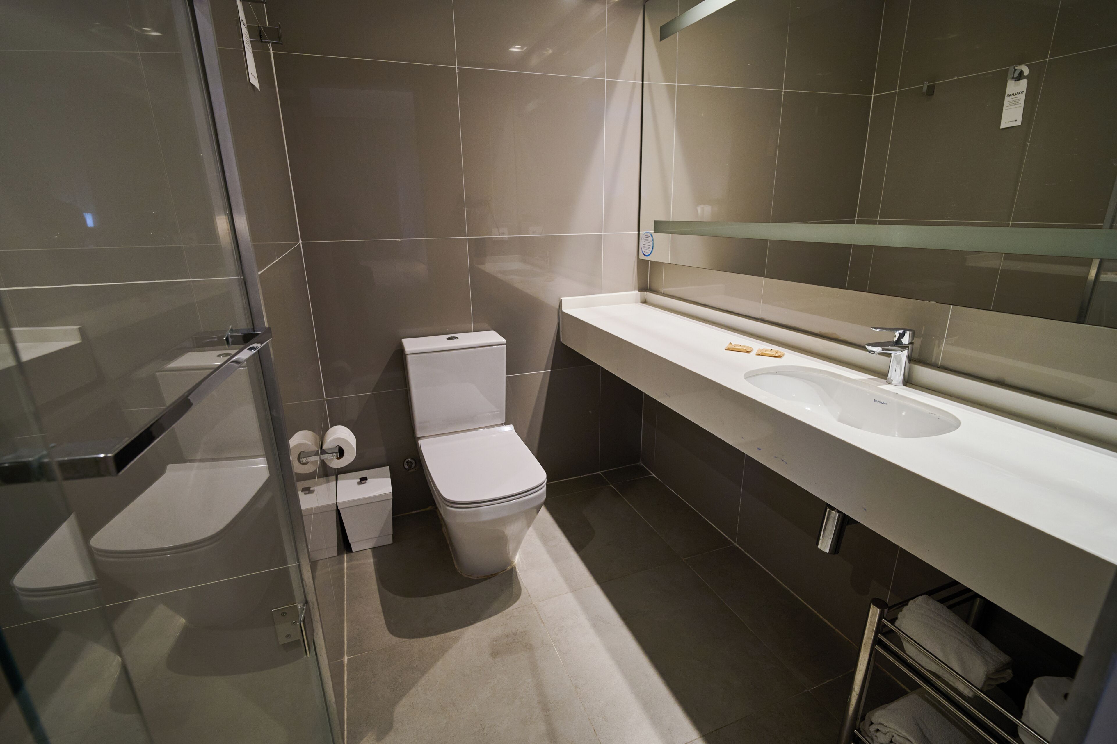 Une salle de bain moderne avec une douche à parois de verre, des toilettes blanches et un grand miroir au-dessus d’un lavabo blanc.