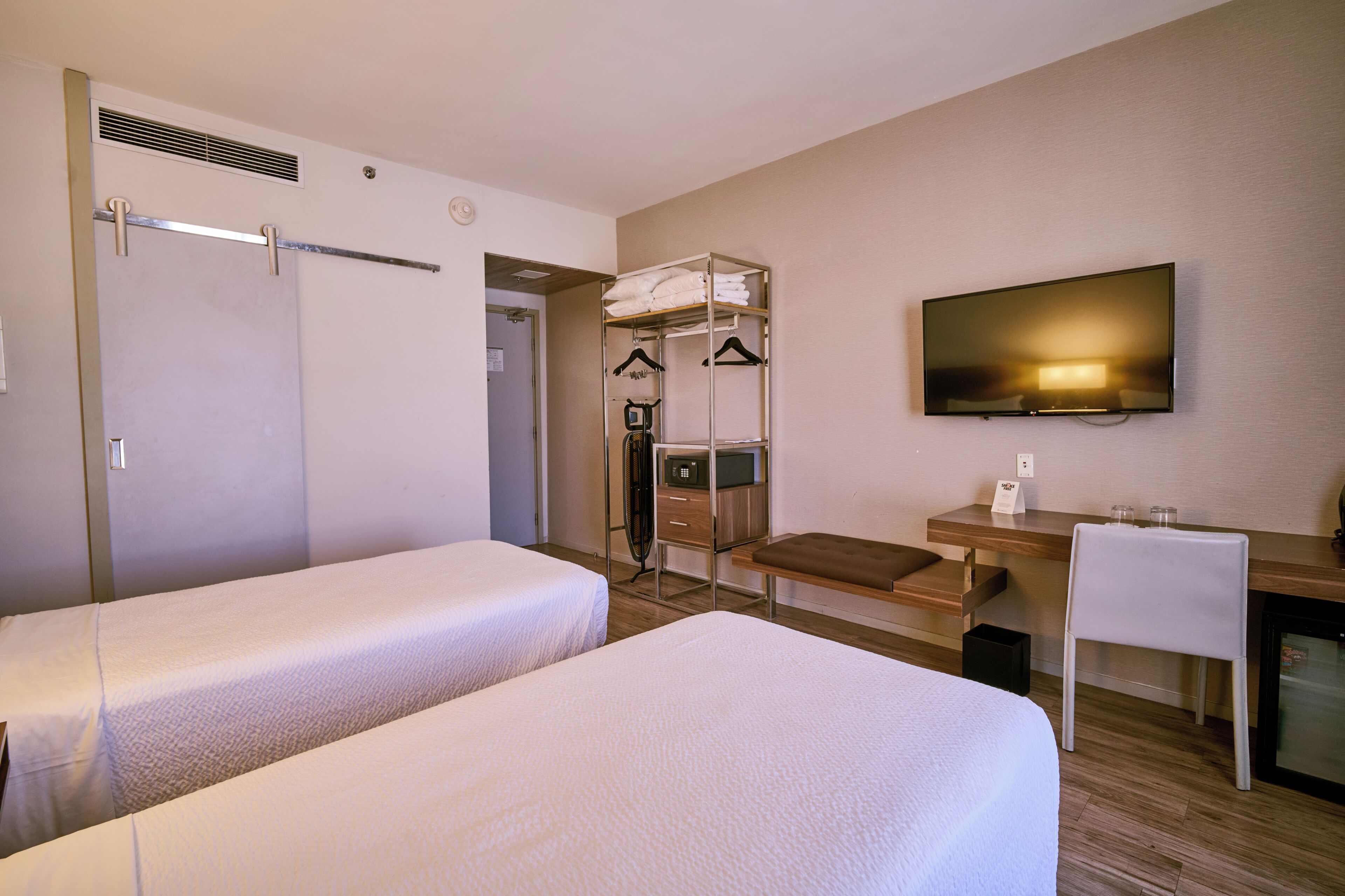 A hotel room with two beds, a desk, a chair, a TV, and a coat rack.